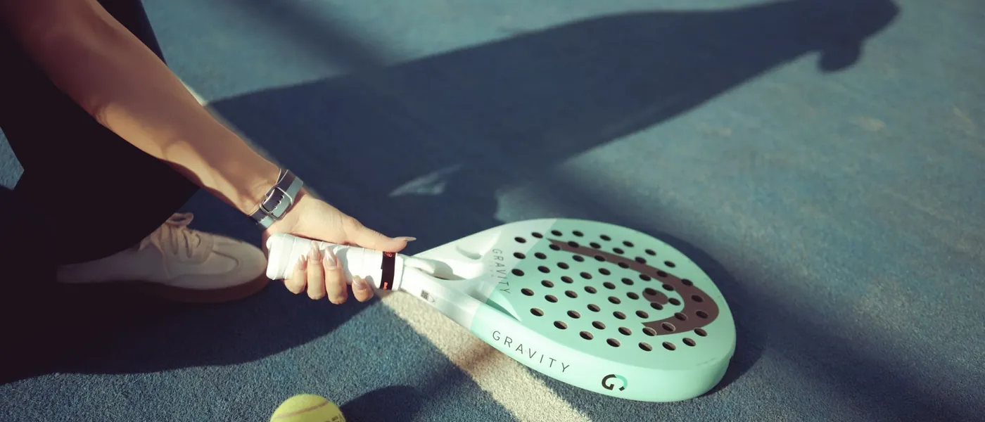 Hand om de greep van een padelracket, met een bal op de baan