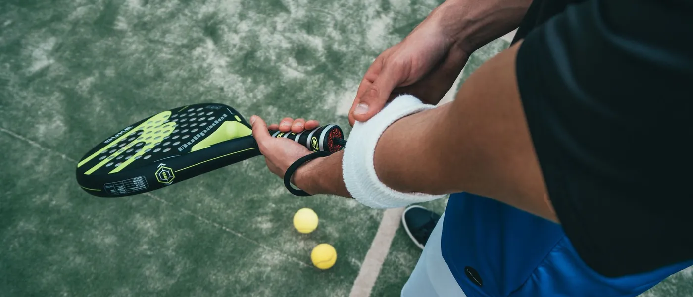 Speler houdt een padelracket vast op de baan, met twee ballen ernaast
